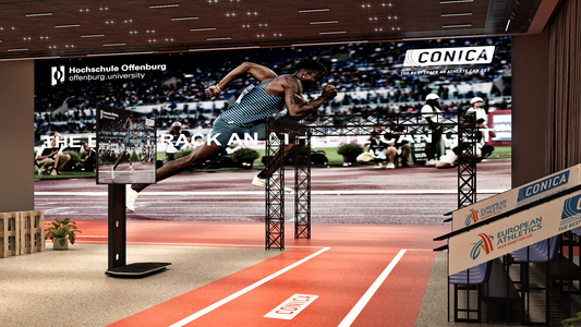 ●	Zu sehen ist das Advanced Motion Laboratory am Campus West. Der Blick ist auf ein 5m x 7m Poster der Firma CONICA gerichtet, welches einen Sprinter zeigt. Davor steht eine schwarze Traverse, an der Infrarotkameras befestigt sind. In den Boden des Labors ist eine 100 m Sprintstrecke aus rotem Tartan eingelassen. Neben der Strecke stehen ein großer Monitor und eine Tribüne für Zuschauer*innen.